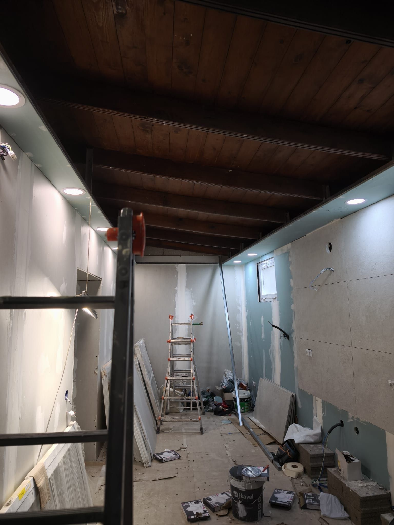 Pasillo estrecho en obras, con una escalera, herramientas, cubos, el techo al descubierto y las paredes sin terminar.