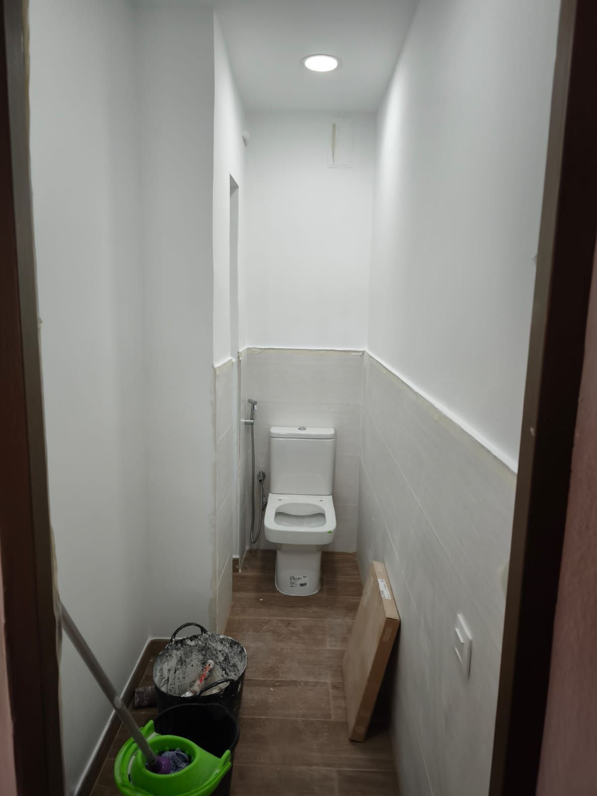Pasillo estrecho con un inodoro instalado al fondo, paredes blancas y productos de limpieza en el suelo.