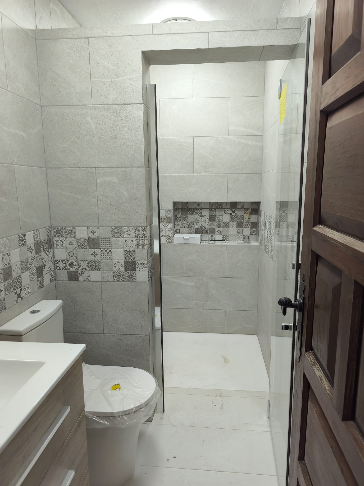 Cuarto de baño pequeño con azulejos, inodoro, lavabo y ducha detrás de una puerta entreabierta.