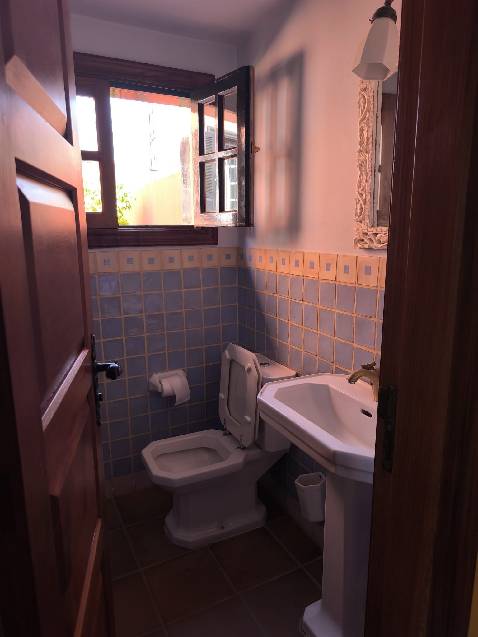 Baño pequeño con inodoro, lavabo, paredes alicatadas y una ventana abierta por la que entra la luz del día.