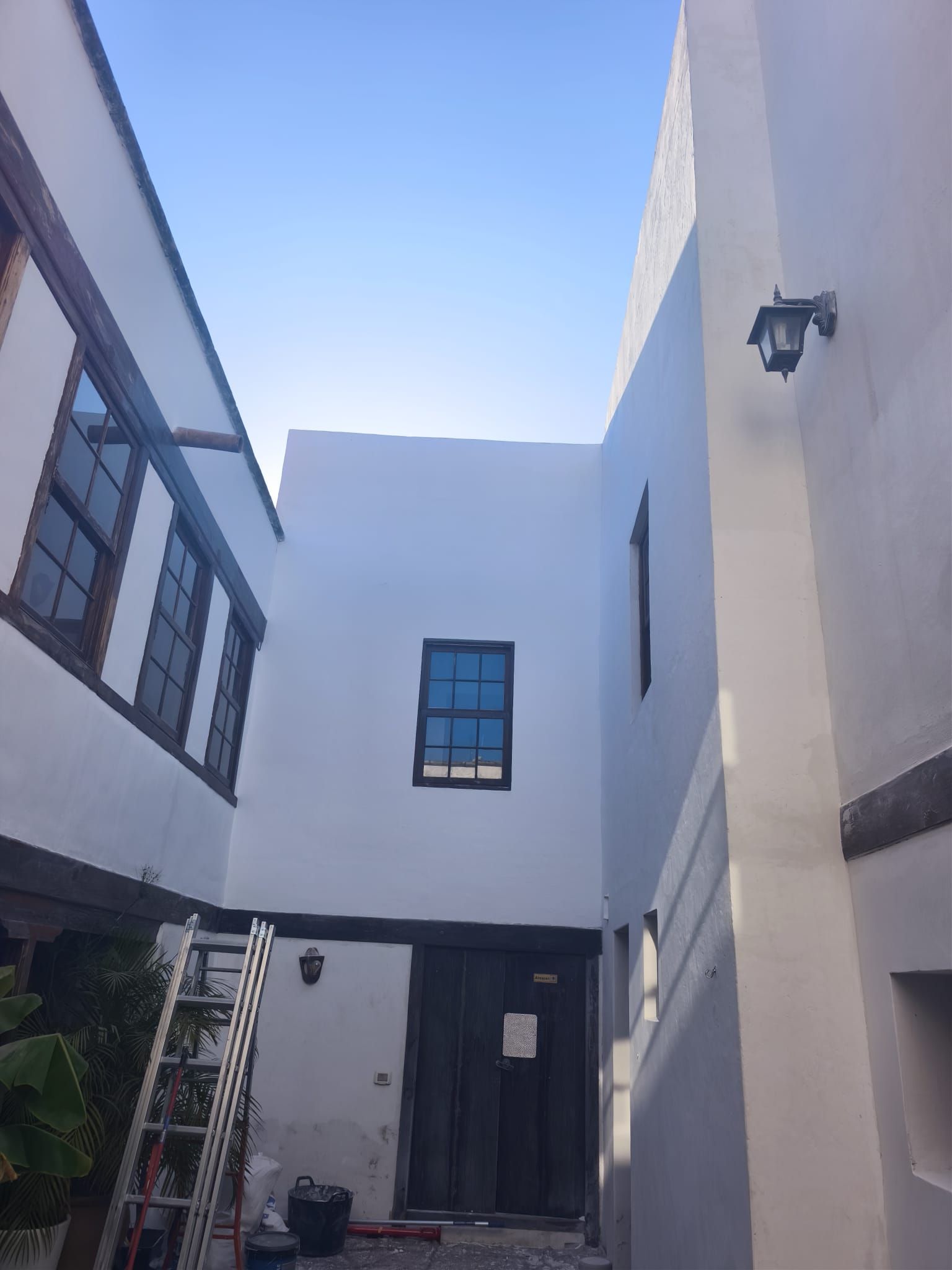 Patio estrecho y blanco, con paredes de dos plantas, ventanas, una puerta negra y una lámpara de pared.