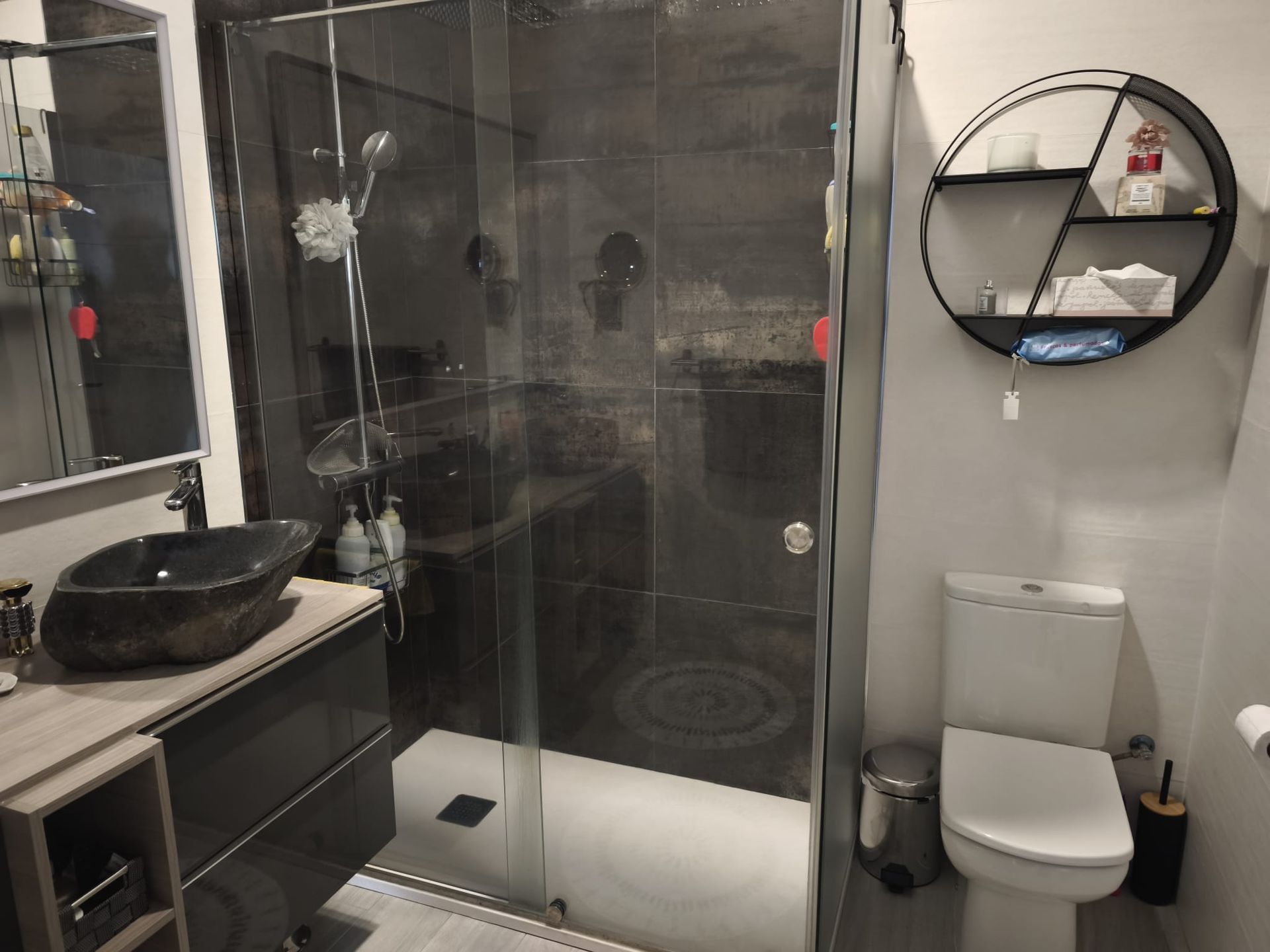 Baño moderno con ducha de cristal, lavabo con mueble y retrete bajo un espejo redondo de pared.