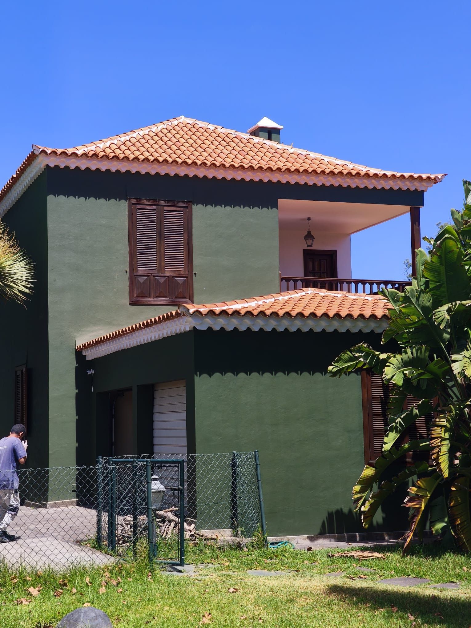 Casa verde de dos plantas con tejado de tejas rojas y balcón, con una persona caminando a su lado bajo un cielo azul.