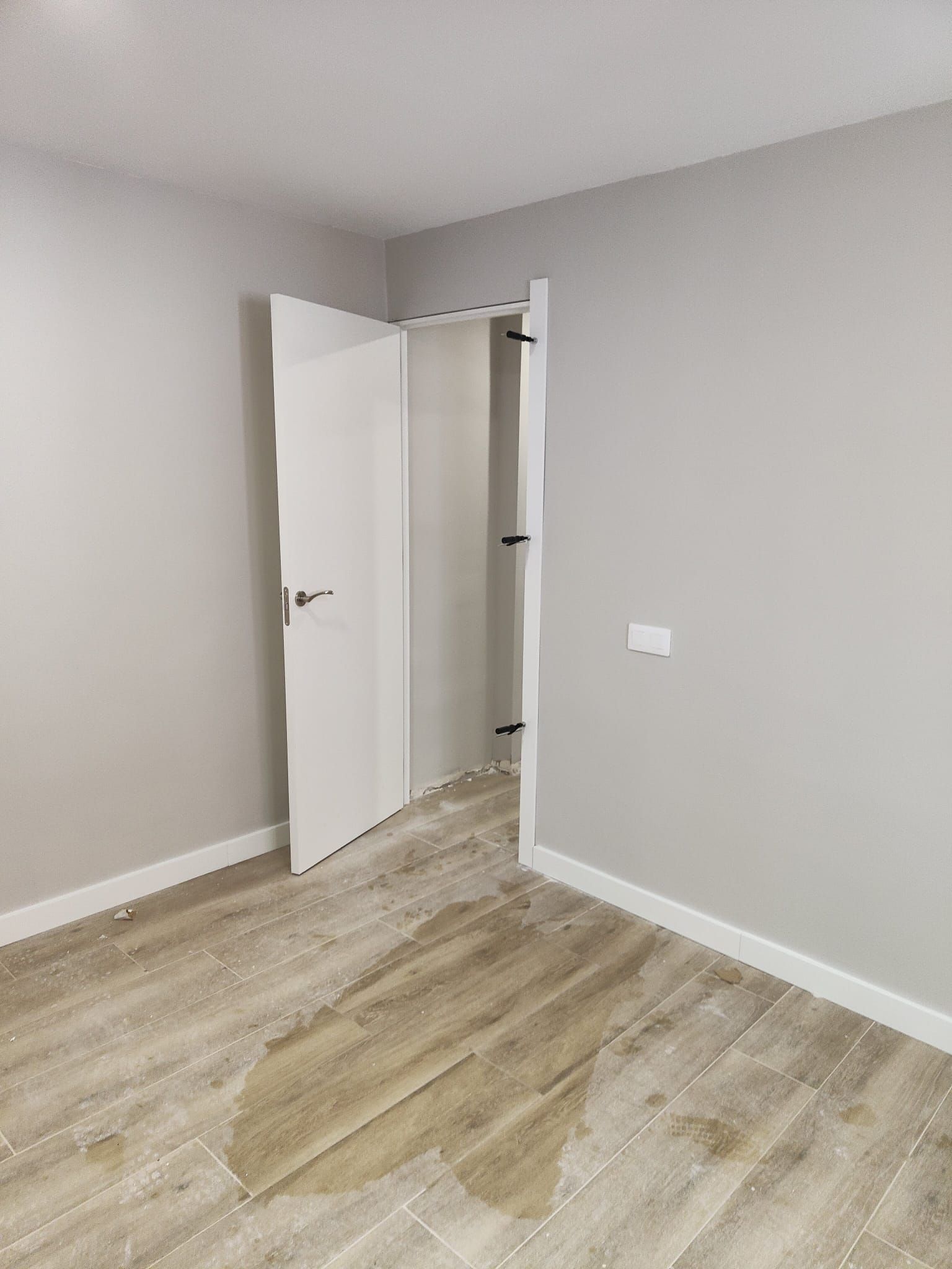 Habitación gris vacía con suelo de aspecto madera y una puerta blanca abierta.