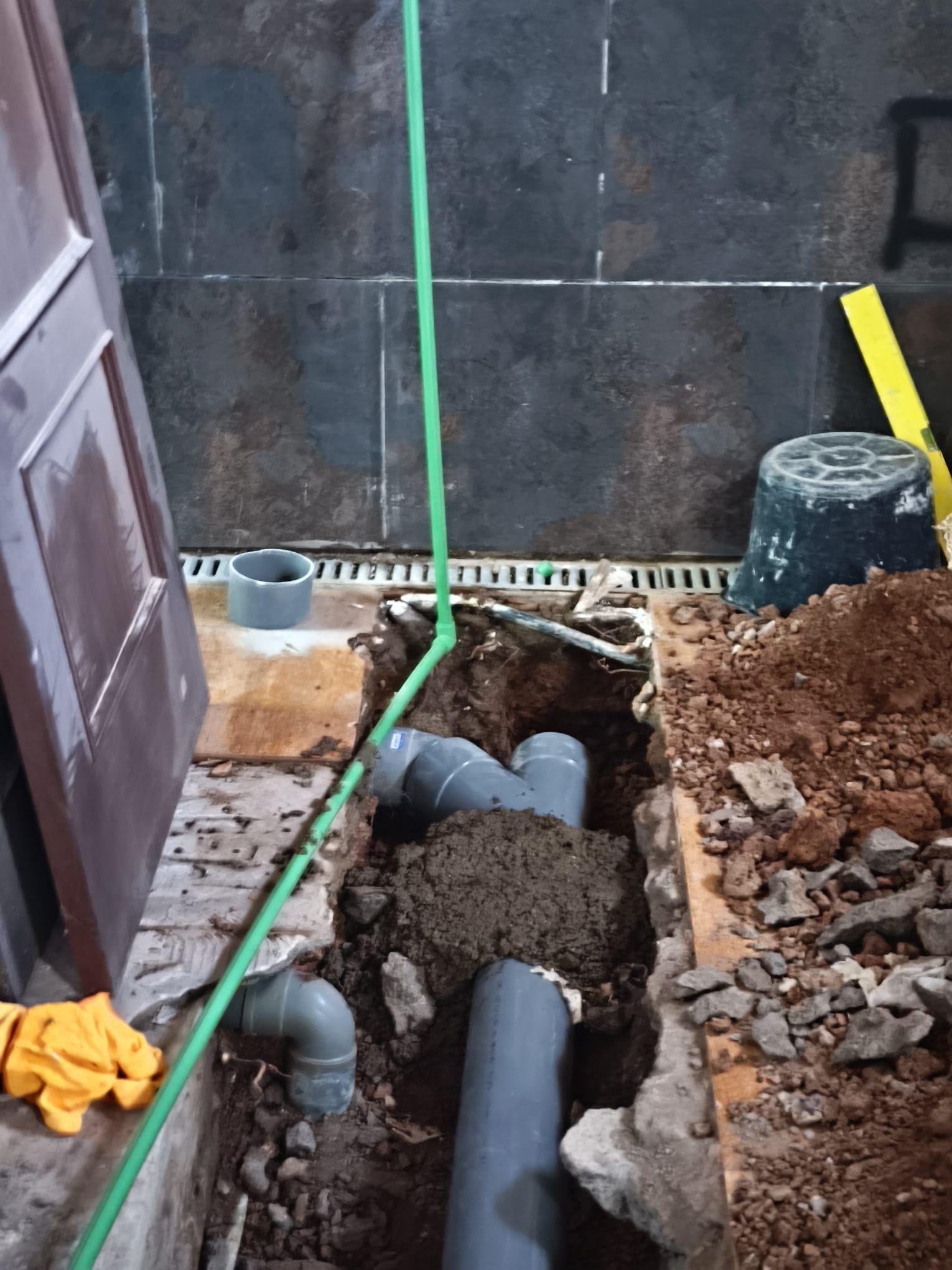 Zanja con tuberías expuestas y hormigón recién vertido junto a una pared, señalizada con una línea verde y cinta amarilla.
