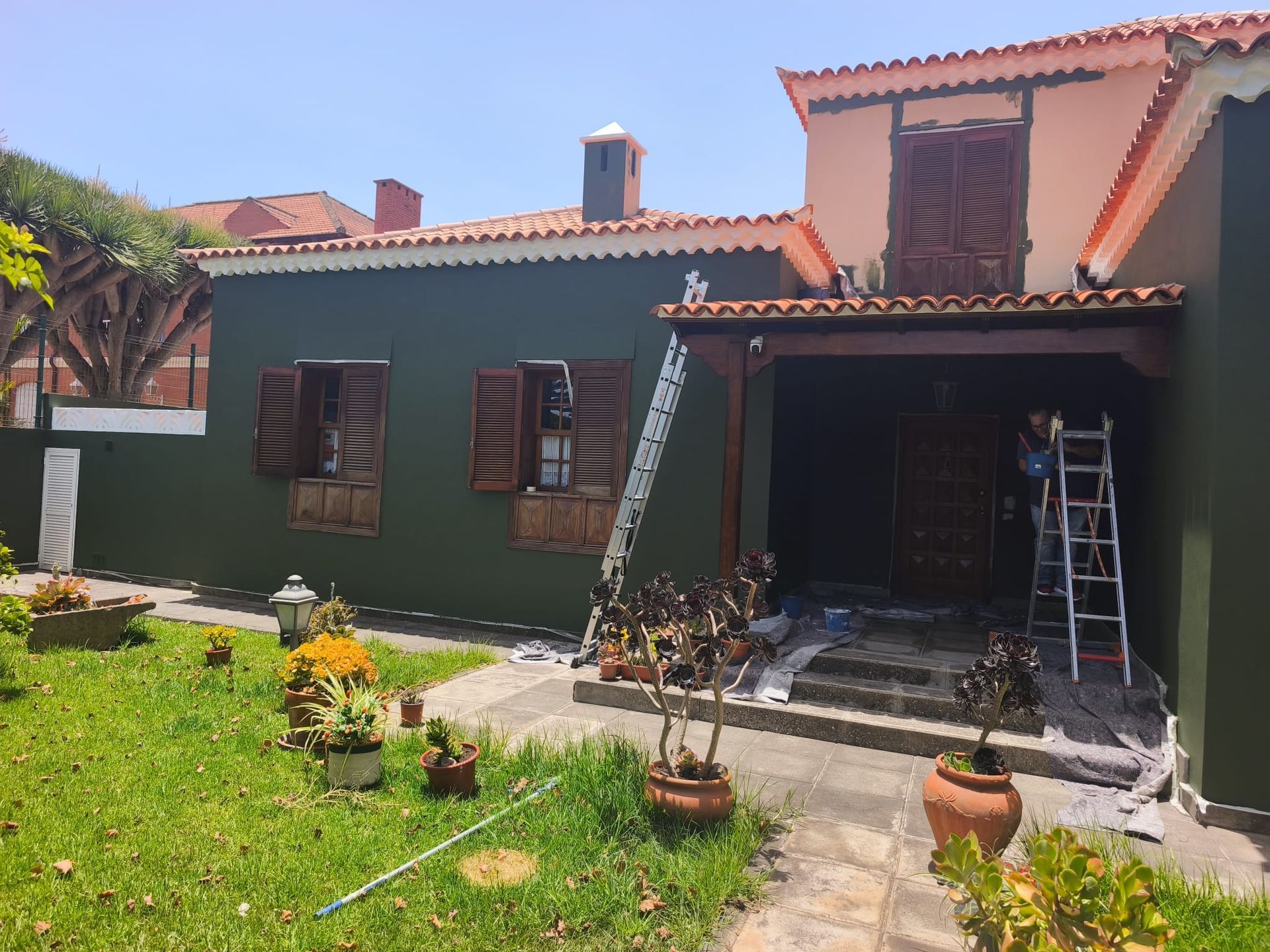 Casa verde con techo de terracota, patio delantero y una escalera junto a la entrada en proceso de renovación.