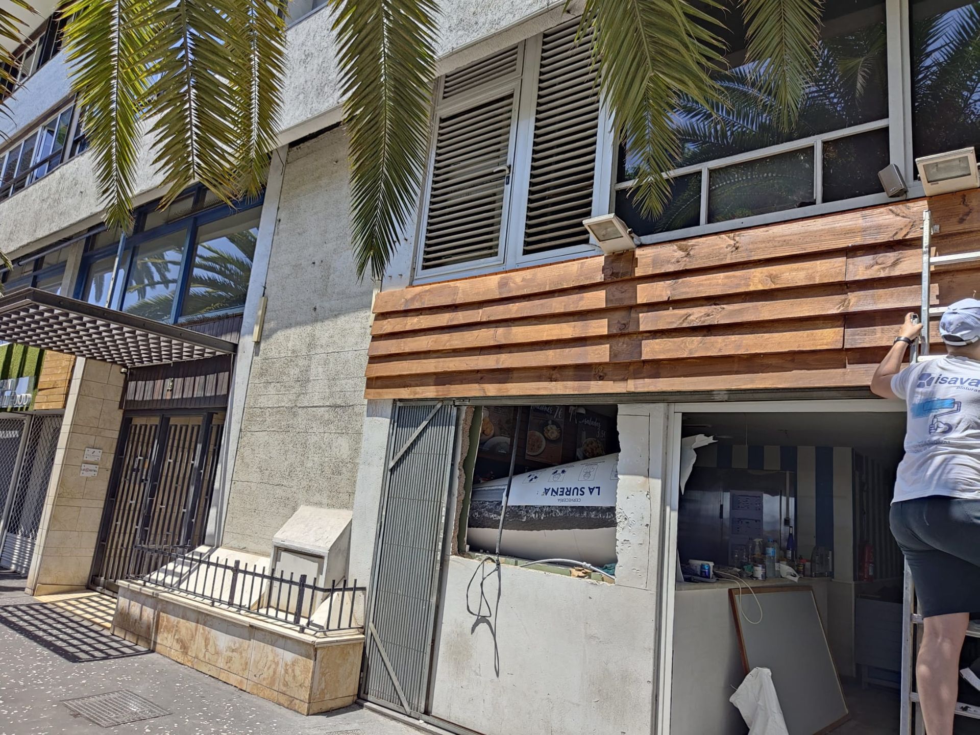 Una persona trepa por el exterior de un edificio, junto a ventanas, revestimiento de madera y una verja metálica, bajo las hojas de una palmera