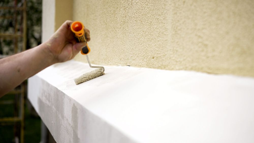 Una mano aplica pintura blanca con un rodillo a lo largo de un saliente exterior junto a una pared beige.