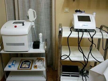 Sala de spa médico con dos máquinas de tratamiento láser blancas sobre carros con ruedas y un estante con suministros.