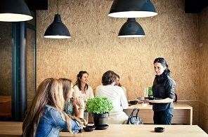 Un camarero se acerca a una mesa donde hay gente sentada en una cafetería con paredes revestidas de madera y lámparas colgantes tipo cúpula.