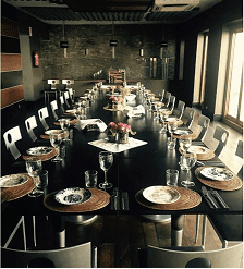 Una mesa de comedor rectangular, larga y oscura, puesta para un evento formal en una sala moderna con paredes de piedra e iluminación industrial.