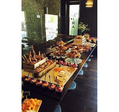 Una larga mesa de banquete puesta con aperitivos, palitos de pan, tazas pequeñas y queso en una habitación rústica con paredes de piedra.