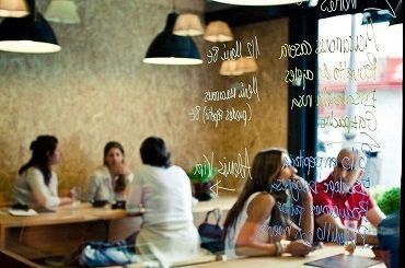 La gente se sienta en mesas en una cafetería bien iluminada, vista a través de una pared de cristal con el menú escrito a mano.