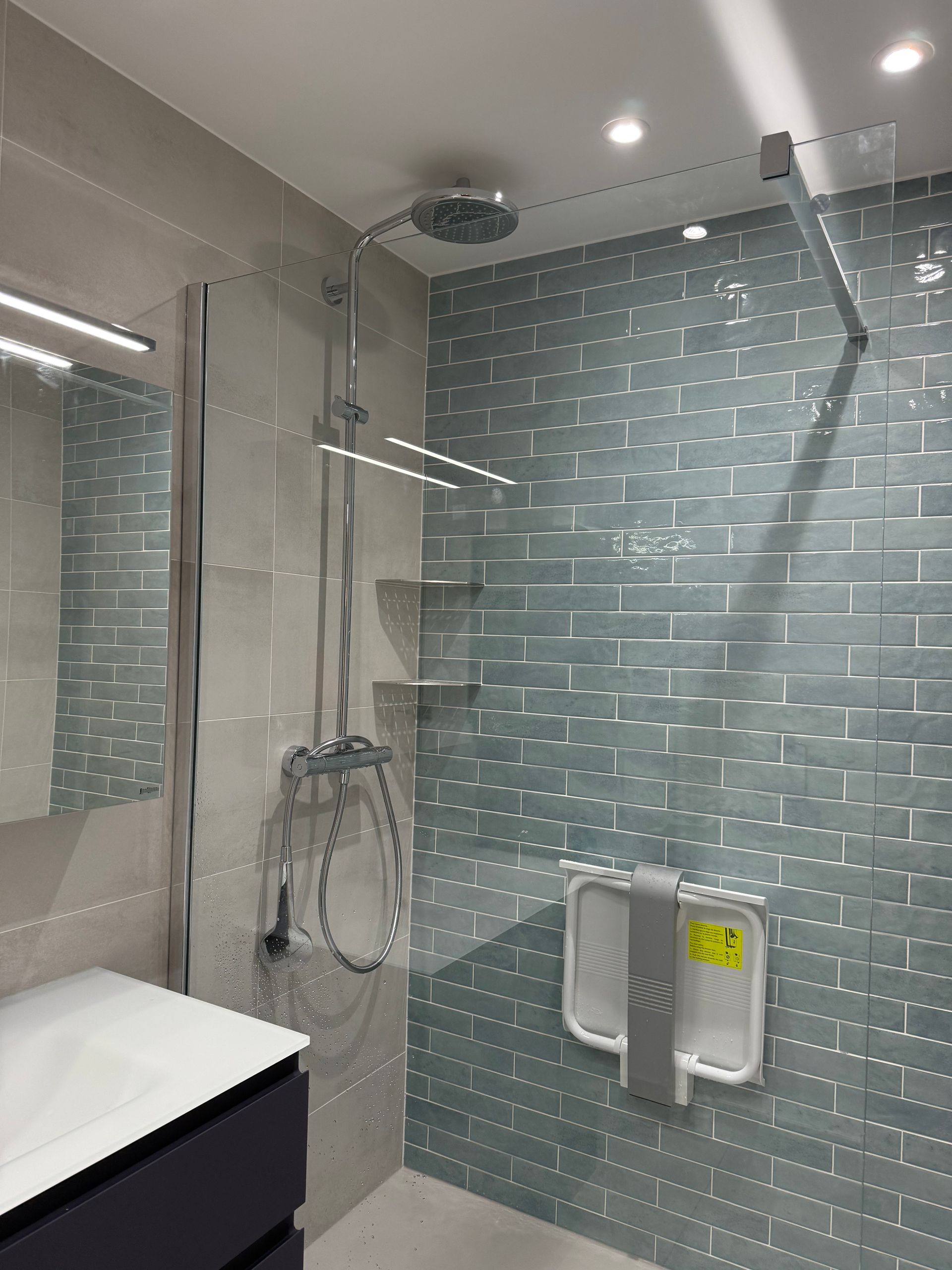 Salle de bains accessible avec douche. Carrelage gris, bleu sarcelle et blanc ; paroi de douche ; robinetterie chromée.
