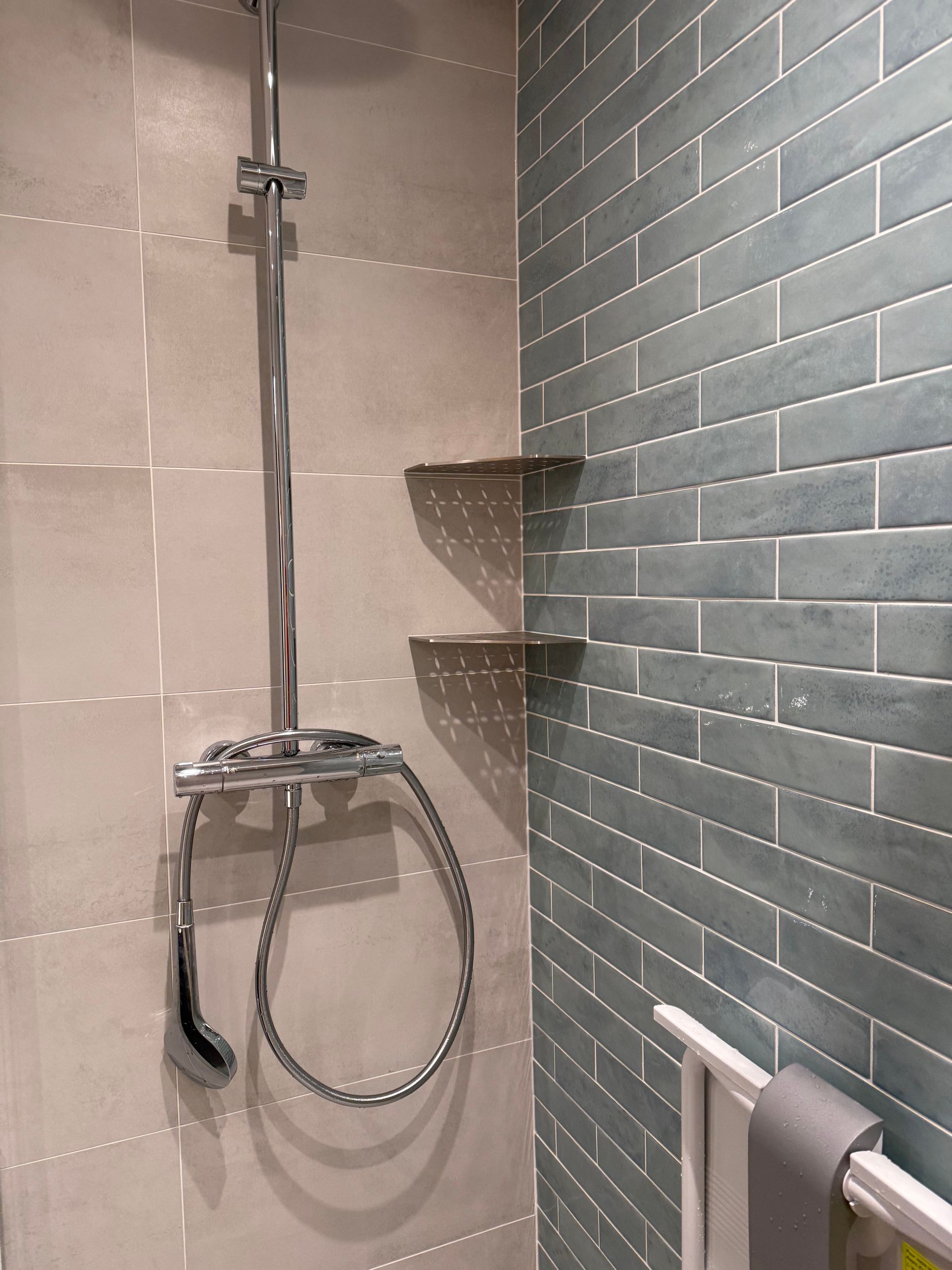 Douche avec robinetterie argentée, carrelage bleu sarcelle imitation brique et carrelage gris. Deux étagères sont visibles.