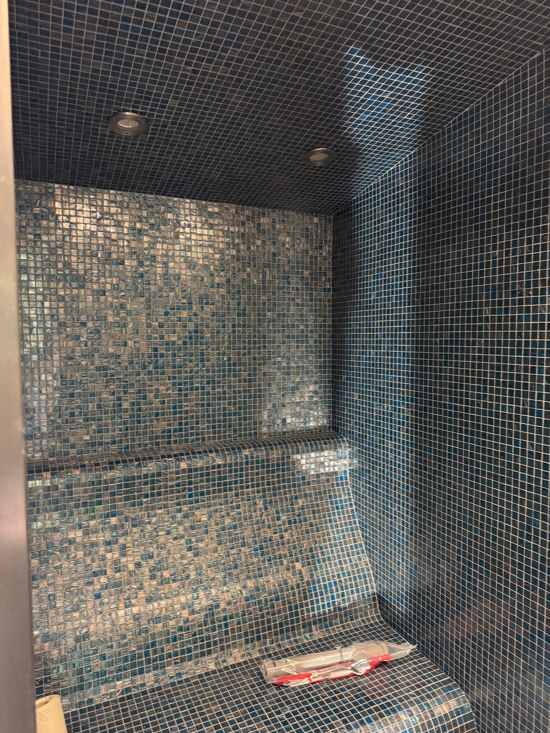 Petite salle de douche carrelée de mosaïques bleues et argentées. Un siège est visible.