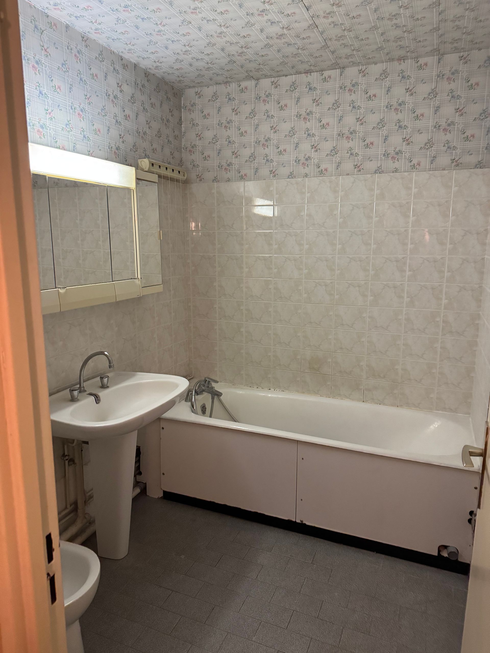 Salle de bain avec lavabo sur colonne, baignoire et papier peint fleuri.