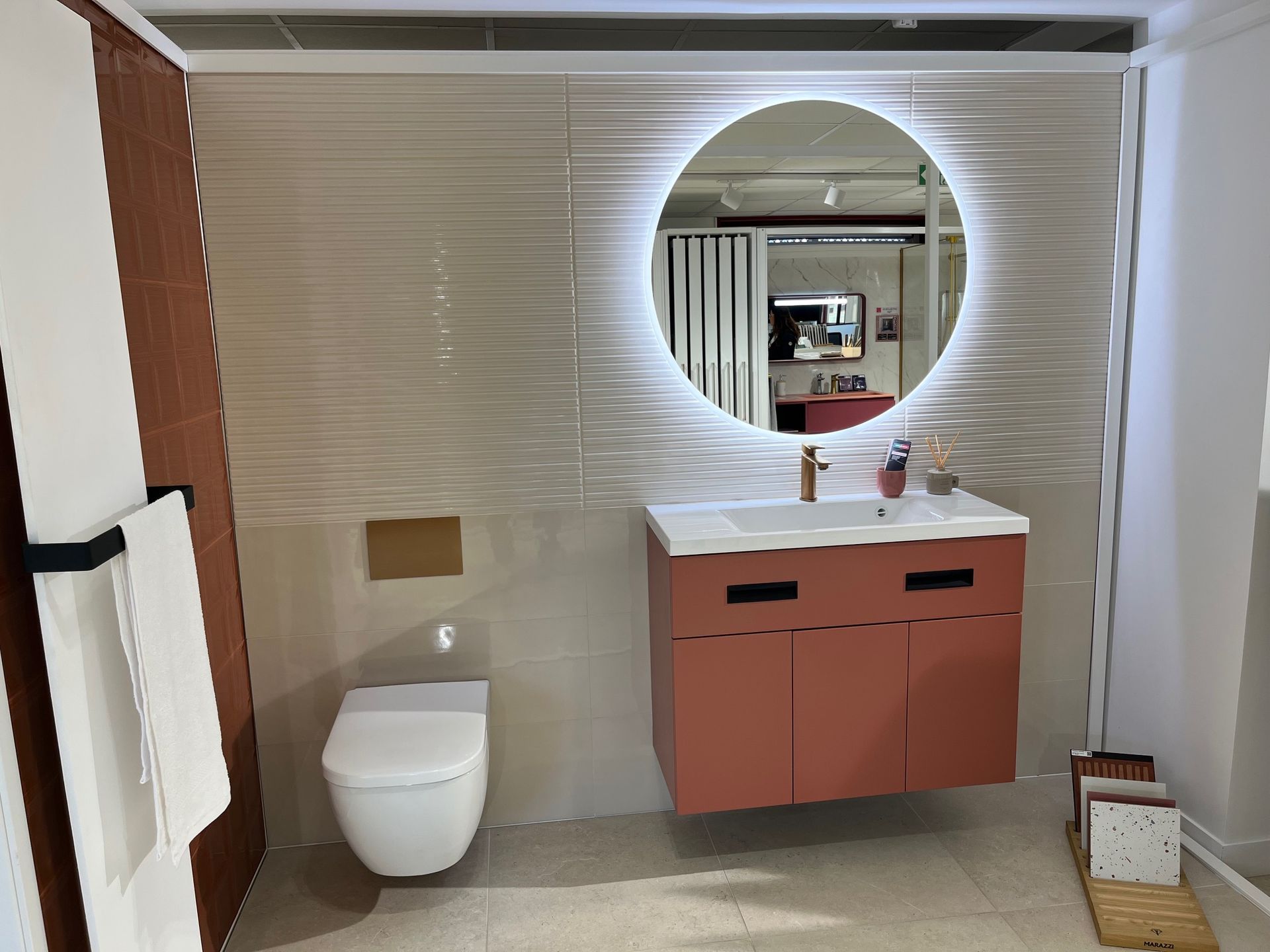 Salle de bain avec armoire et mur d'accent en terre cuite, miroir rond éclairé et toilettes blanches.