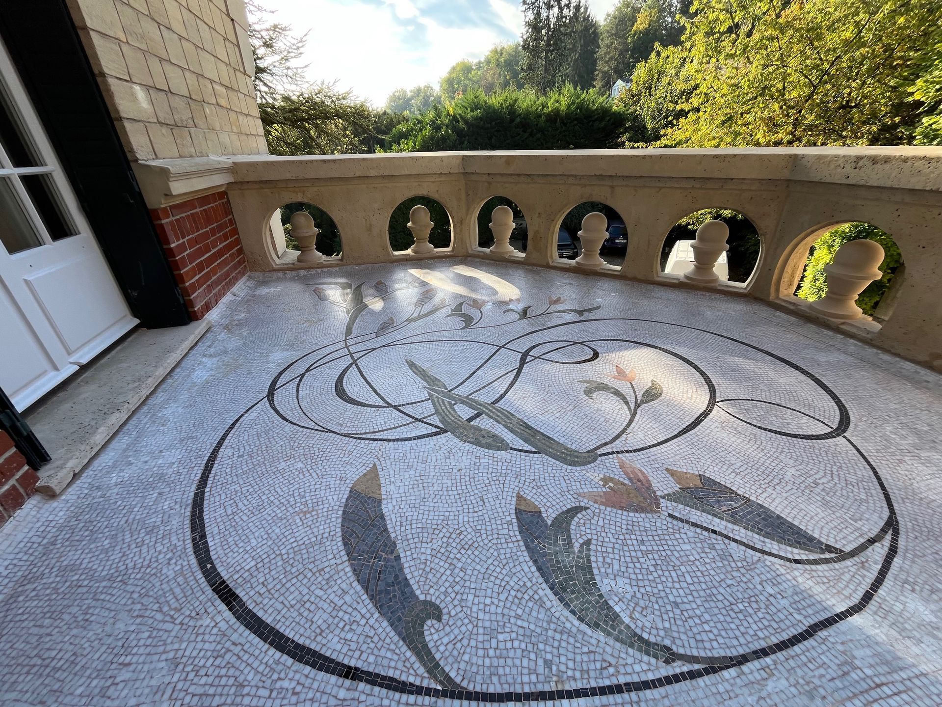 Balcon avec sol en mosaïque, balustrade en pierre ornée et arbres en arrière-plan.
