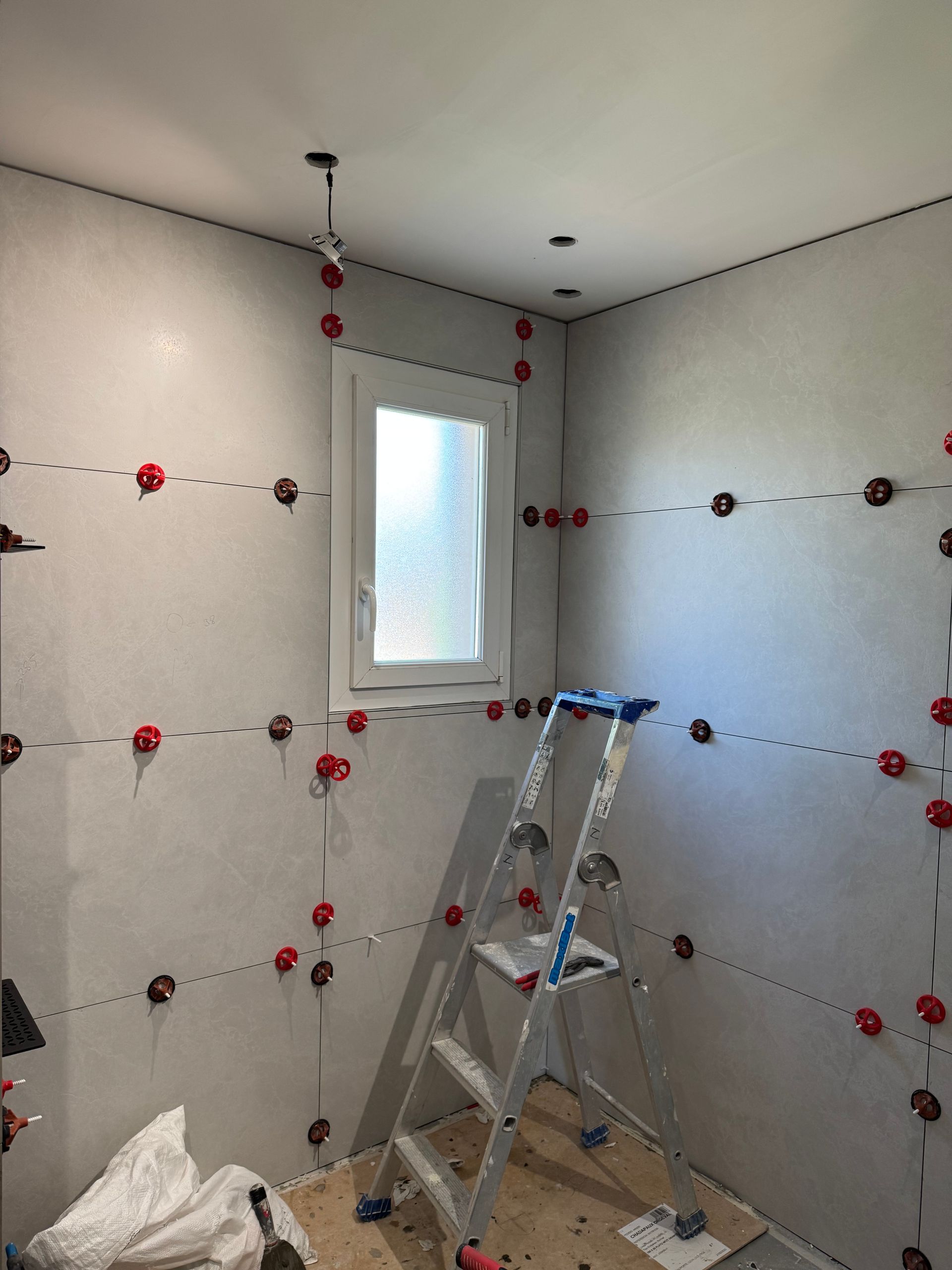 Pièce intérieure en construction; murs gris avec entretoises de nivellement de carrelage rouge; escabeau près de la fenêtre.