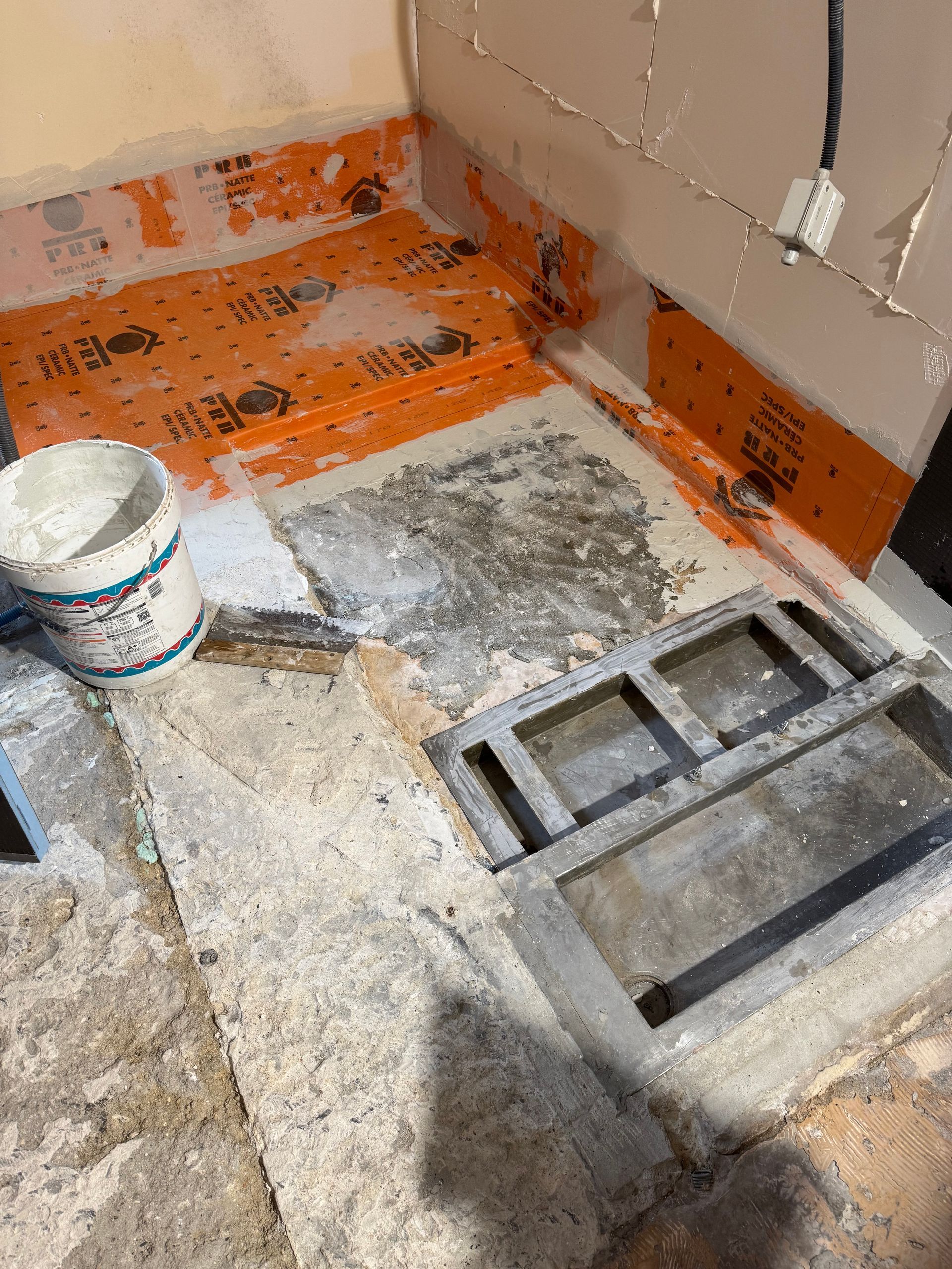 Construction de douche : Membrane imperméable orange installée, base de béton, drain gris, seau de coulis.