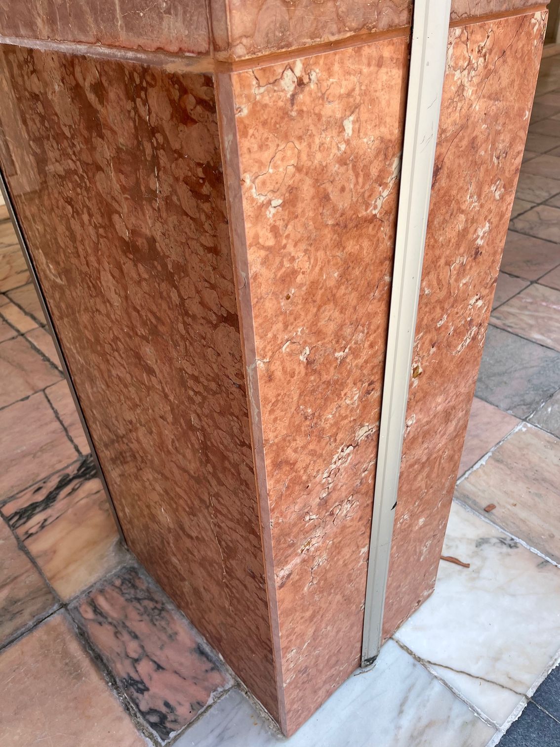 Colonne en pierre rouge et brune à cerclage clair, posée sur un sol en marbre aux veinures contrastées.