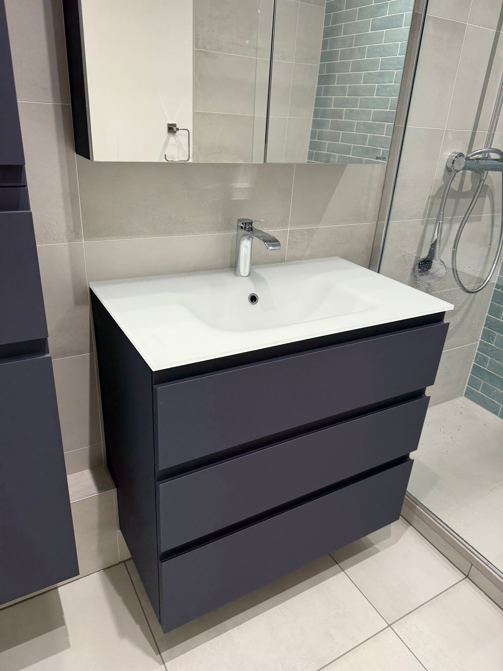 Salle de bain avec vanité bleu foncé, lavabo blanc et miroir; carrelage clair, douche.