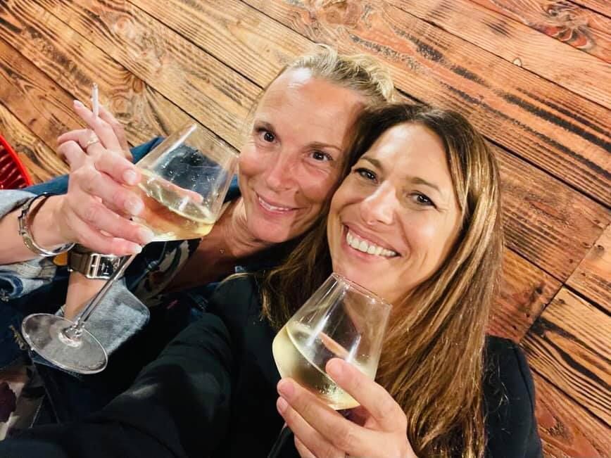 Deux femmes souriantes, trinquant avec des verres de vin, fond en bois.
