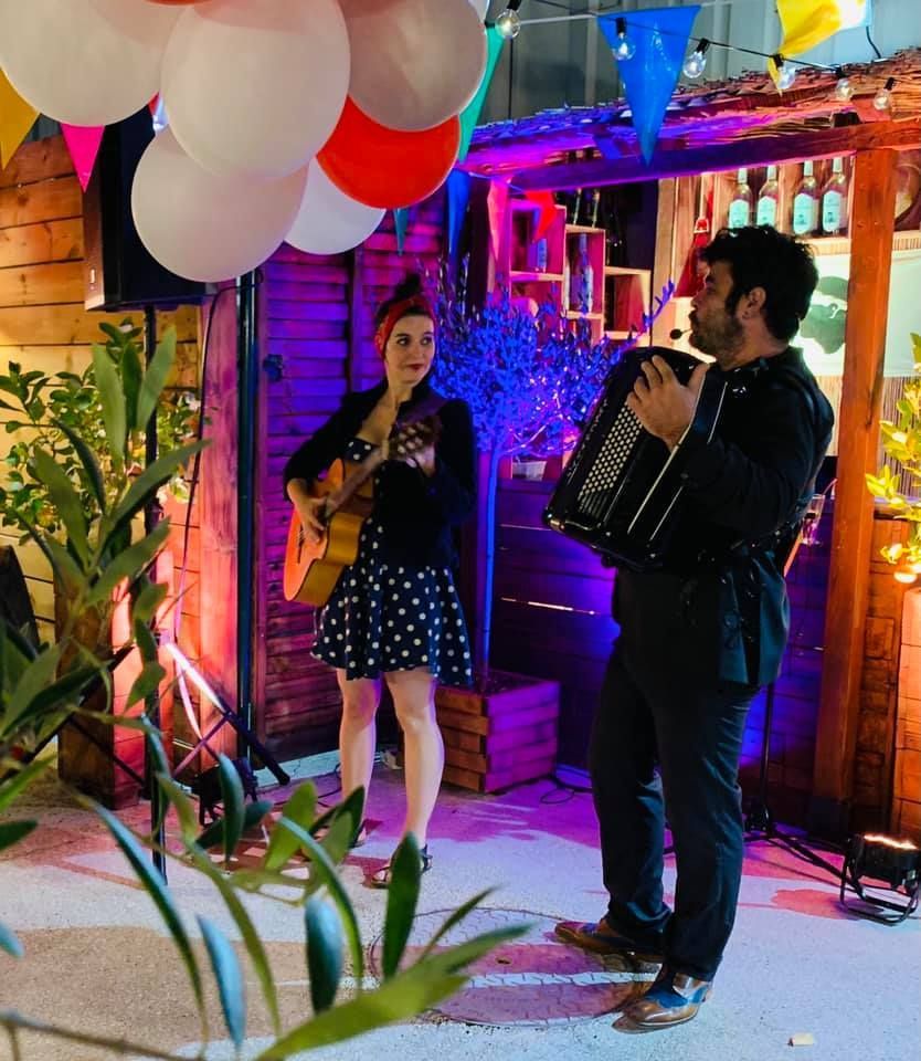 Des musiciens jouent en plein air : une femme avec une guitare, un homme avec un accordéon, des décorations colorées.