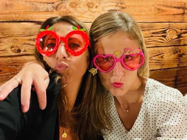 Deux femmes posent, portant des lunettes de soleil fantaisie et faisant des grimaces de baiser. Fond en bois.