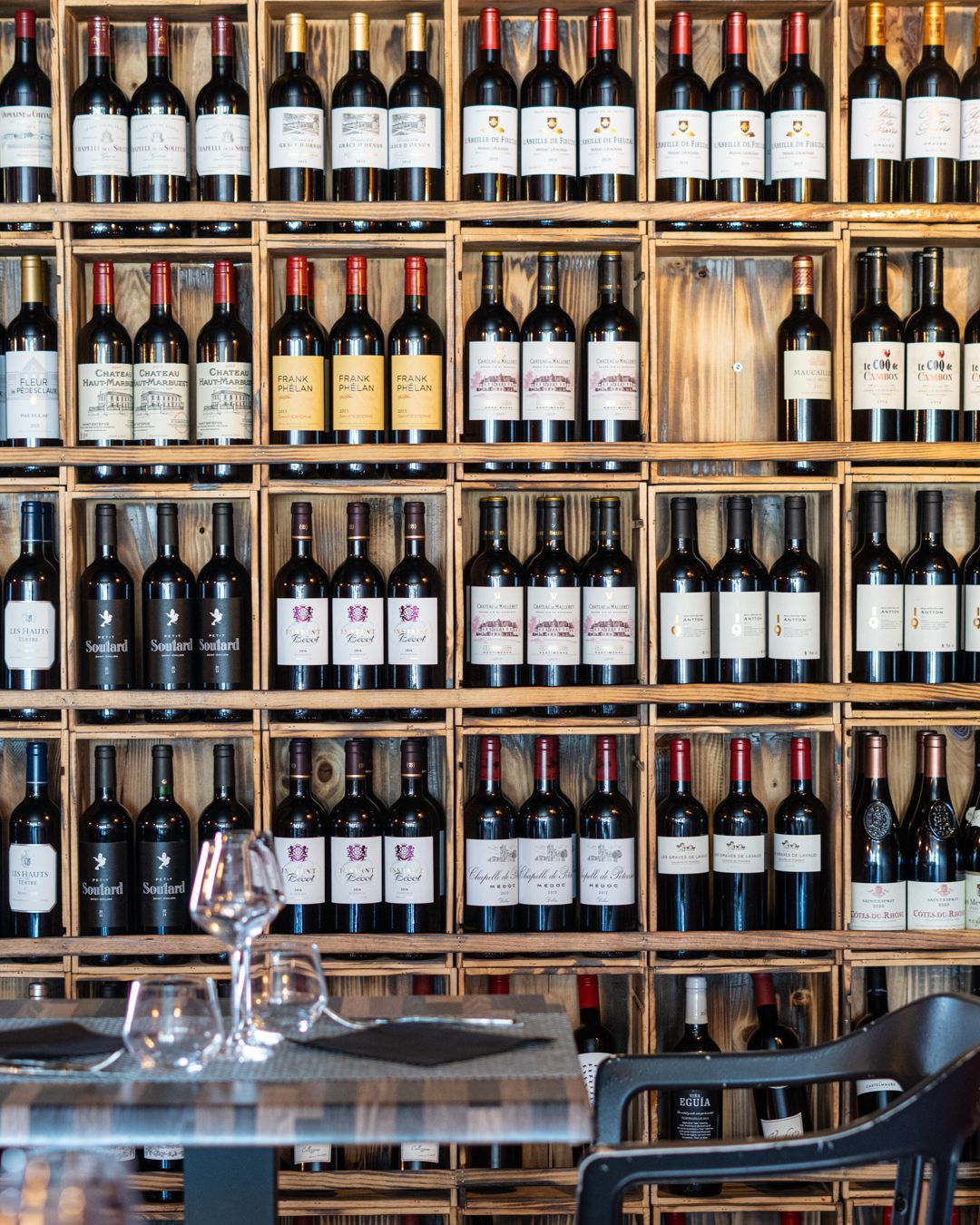Des bouteilles de vin sont présentées dans des caisses en bois sur un mur, avec une table dressée devant.