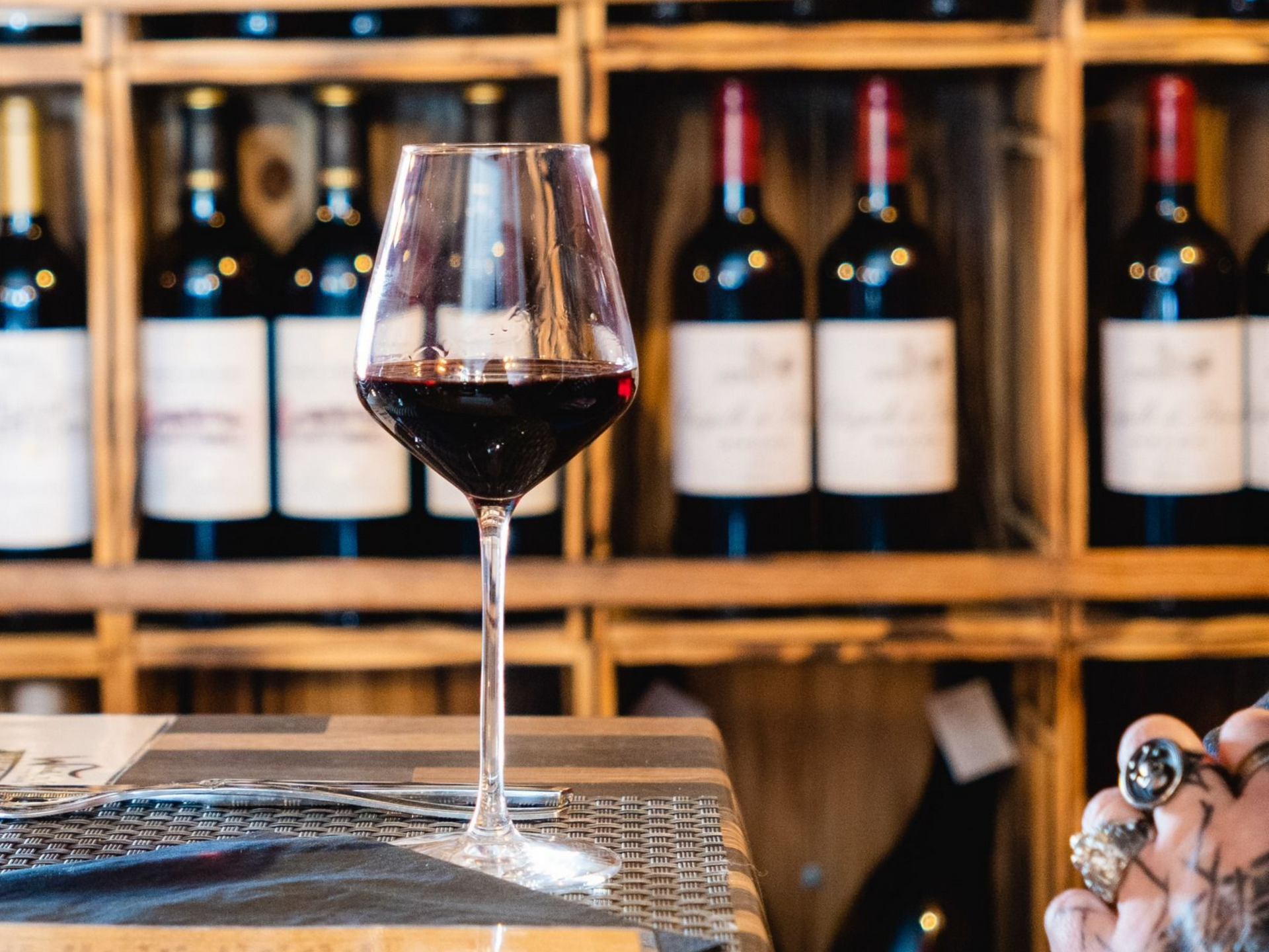 Verre à vin rempli de vin rouge, devant un fond de bouteilles de vin rangées sur des étagères en bois.