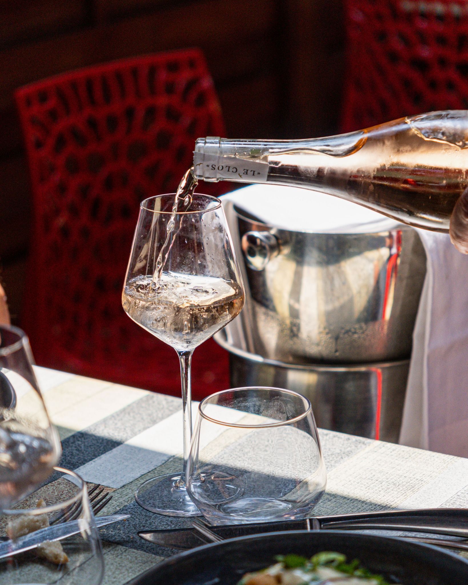 Du vin est versé dans un grand verre dans un restaurant, avec une table dressée et un rafraîchisseur à vin en arrière-plan.