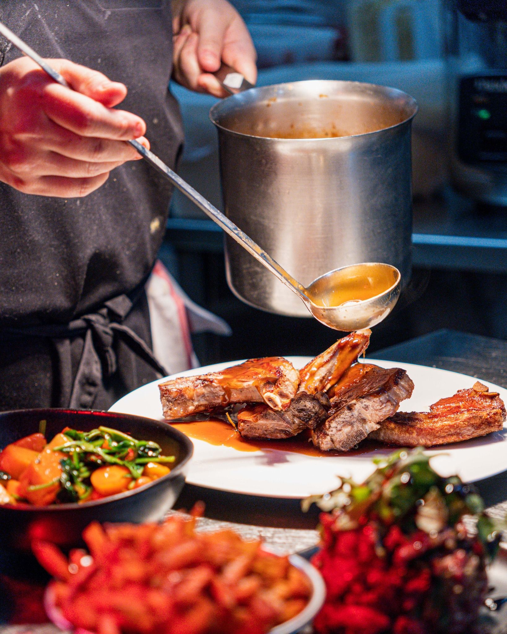 Le chef nappe de sauce une assiette de viande cuite. On aperçoit les accompagnements et les ustensiles de cuisine.