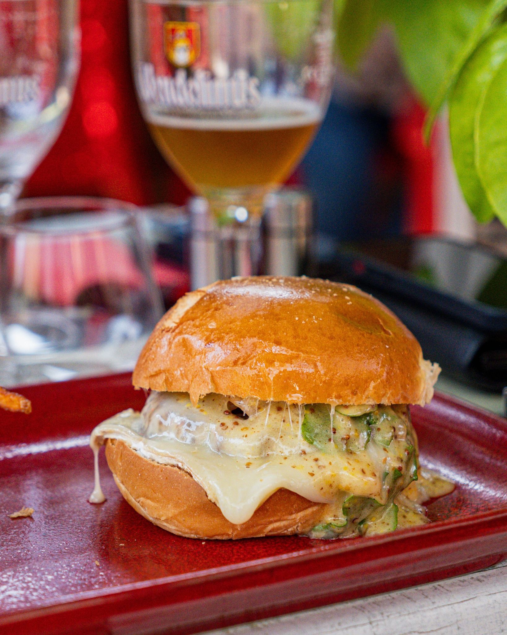 Un hamburger avec du fromage fondu et de la sauce, servi sur une assiette rouge, avec une bière en arrière-plan.