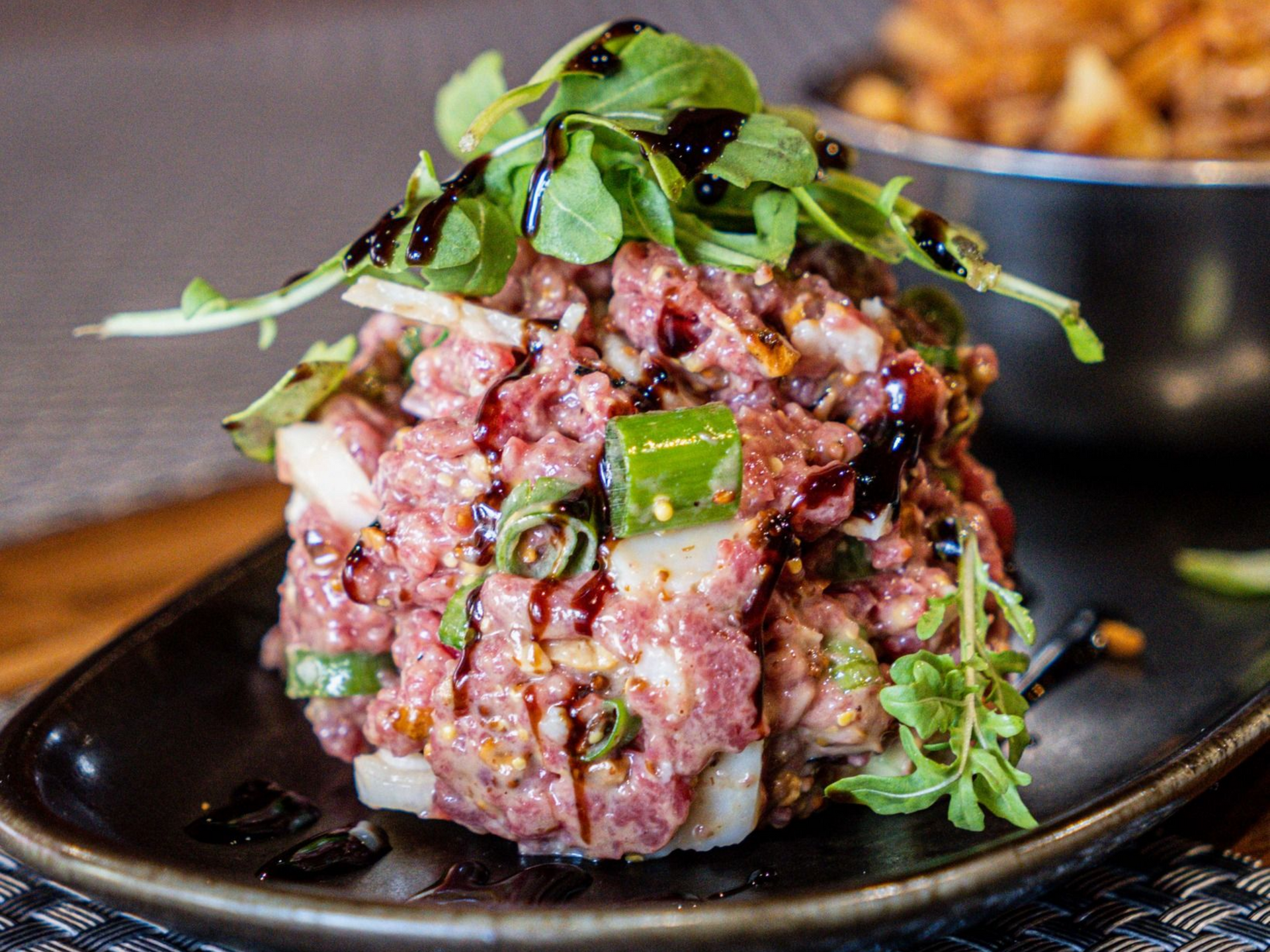 Tartare de bœuf, garni de roquette et d'un glaçage balsamique, servi sur une assiette noire.