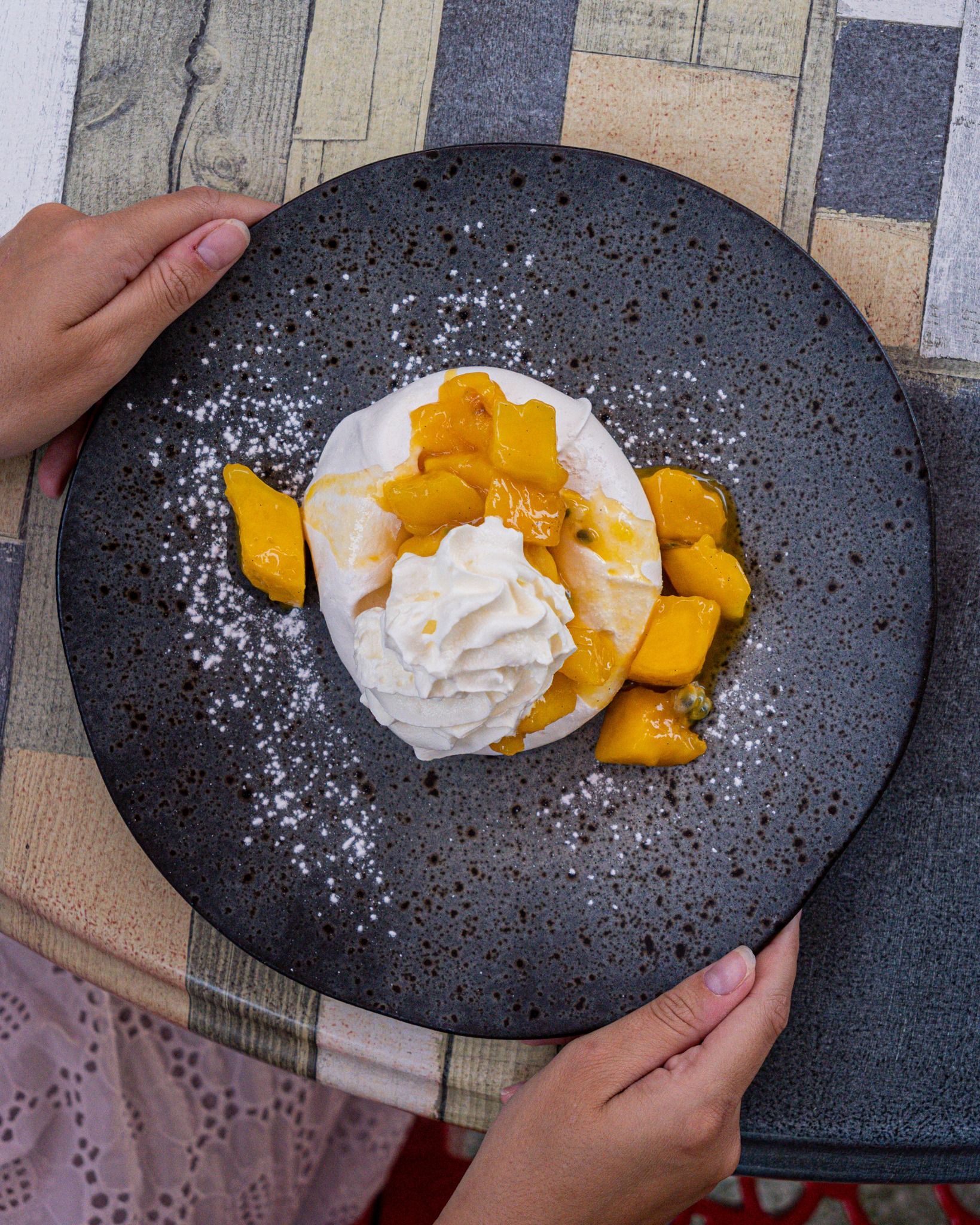 Dessert : meringue avec crème fouettée et morceaux de mangue sur une assiette gris foncé, à déguster à la main.