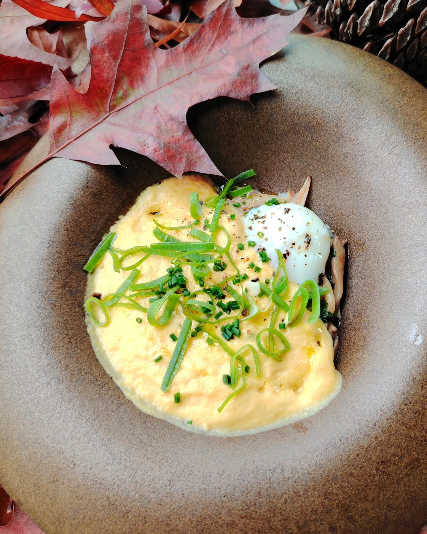 Œufs brouillés aux ciboulettes dans un bol brun, garnis d'un œuf poché. Feuilles d'automne en arrière-plan.