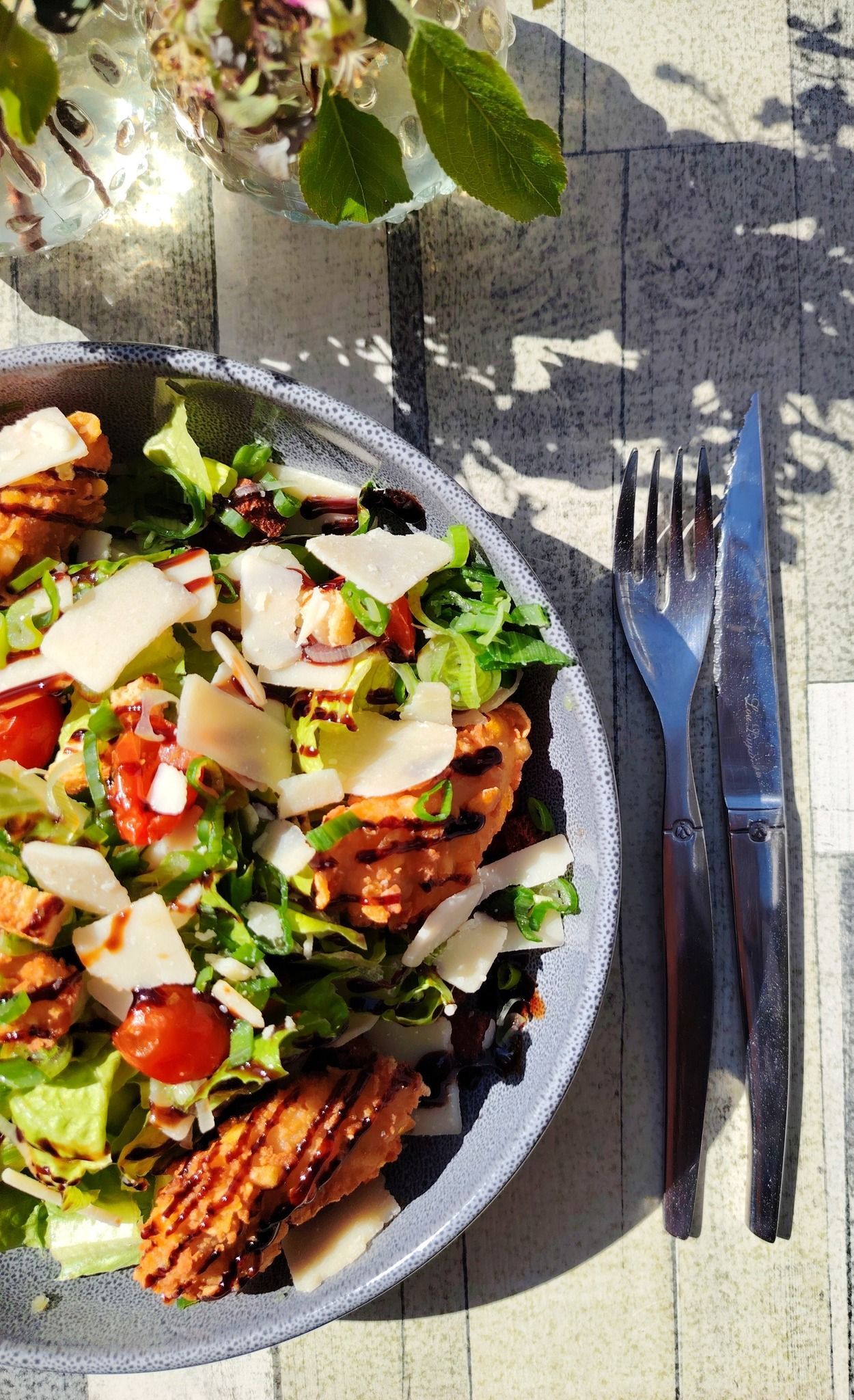 Salade avec poulet frit, tomates et parmesan sur une assiette grise, à côté d'une fourchette et d'un couteau.