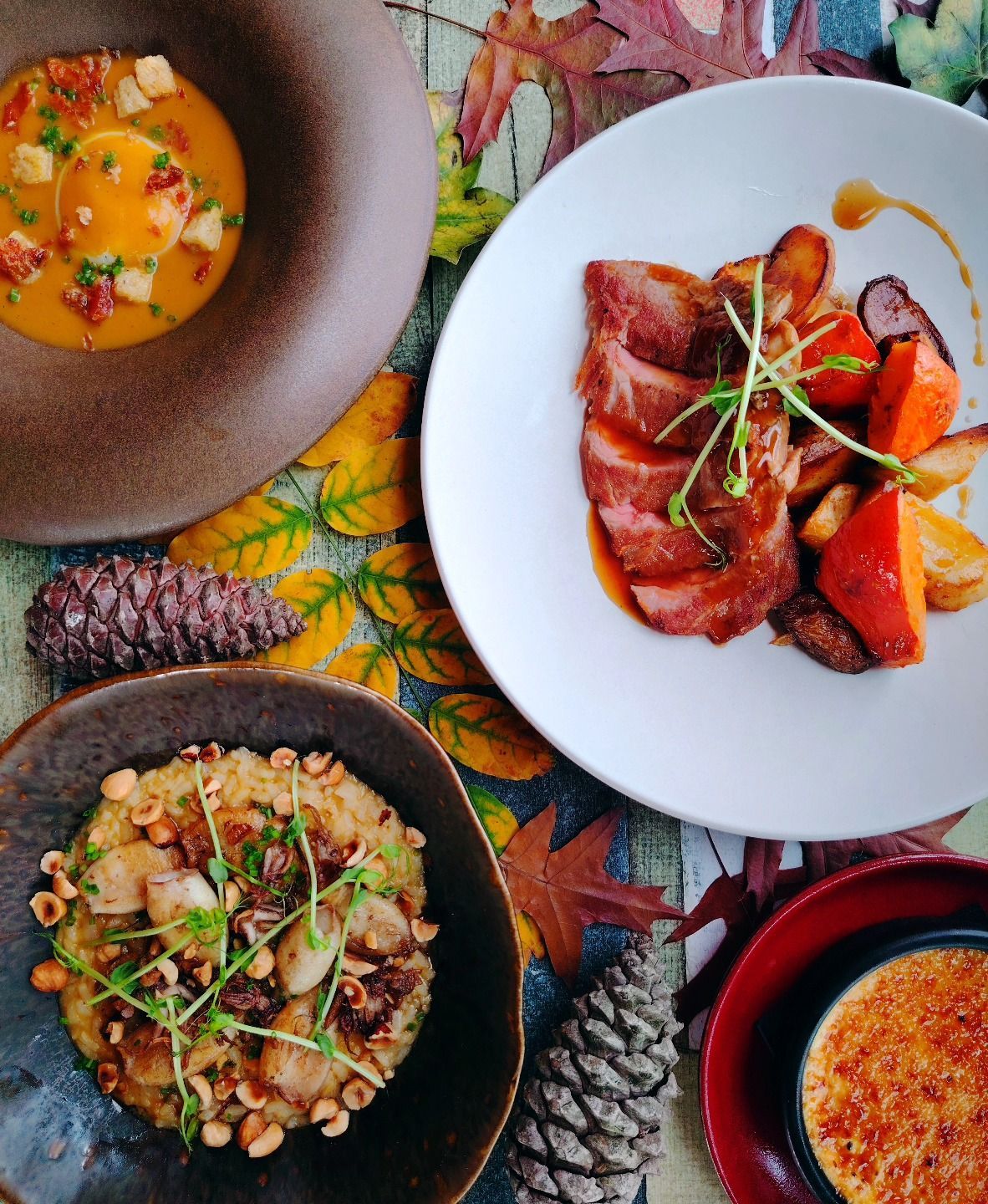 Gros plan sur quatre plats présentés sur une surface aux couleurs d'automne : soupe, viande, risotto et crème brûlée.