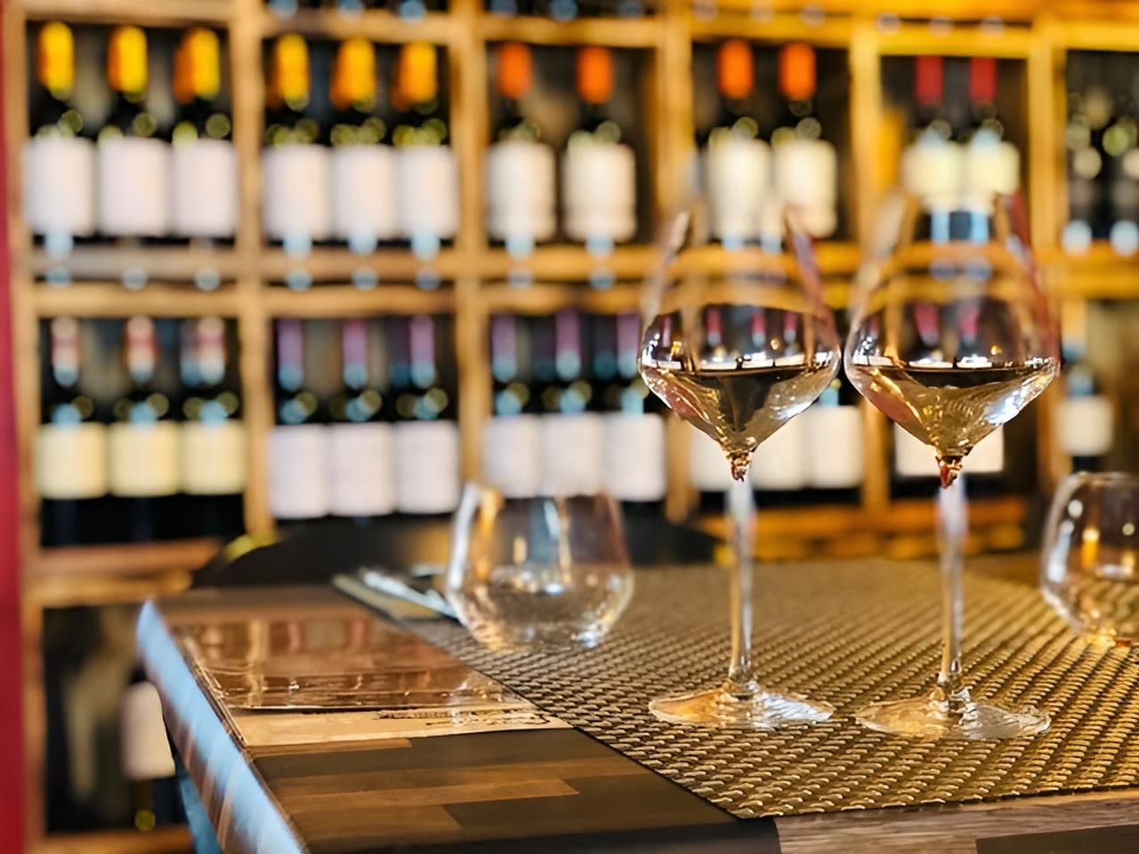 Deux verres à vin remplis de liquide sur une table en bois, des bouteilles de vin en arrière-plan.