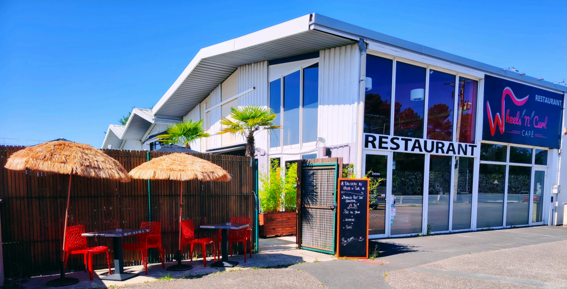 Façade du restaurant avec chaises rouges, parasols en paille et menu sur ardoise. Le bâtiment est blanc avec de grandes fenêtres.