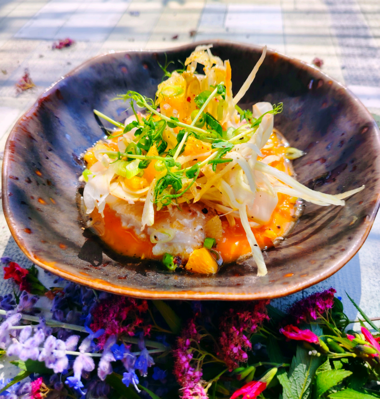 Un plat de fruits de mer gastronomique servi dans un bol en céramique foncée, garni de micro-pousses et disposé parmi des fleurs sauvages colorées.