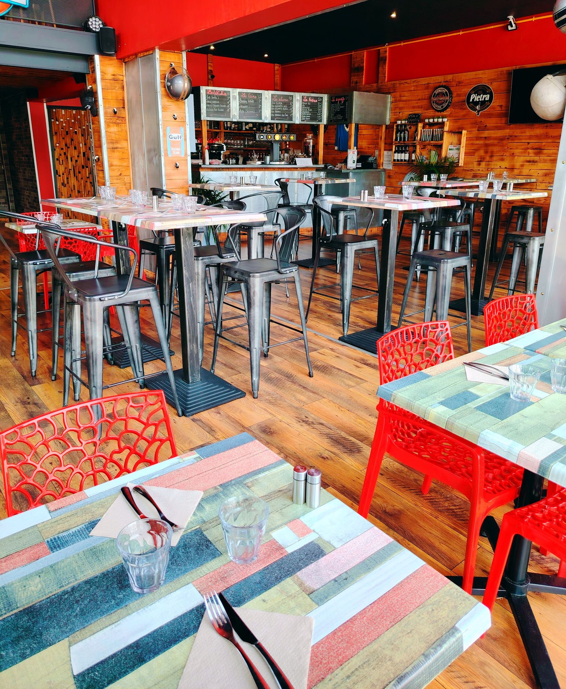 Intérieur du restaurant avec tables hautes, sièges de bar, parquet, tables et chaises rouges et à motifs.