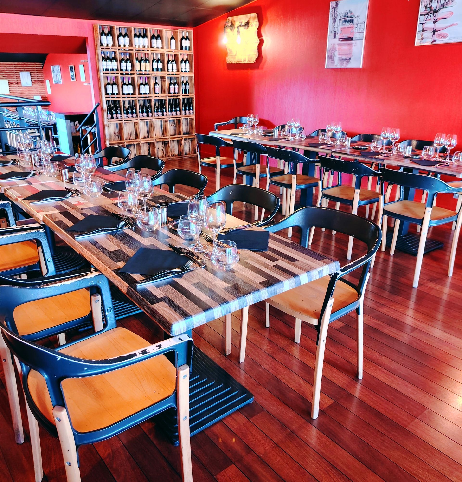 Intérieur d'un restaurant avec une table en bois, des chaises sombres, des murs rouges et un casier en bois rempli de bouteilles de vin.