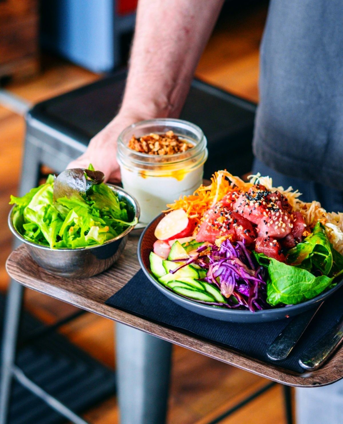 Une personne tenant un plateau en bois avec un grand bol de poke bowl, une salade verte en accompagnement et un petit pot de dessert à étages.