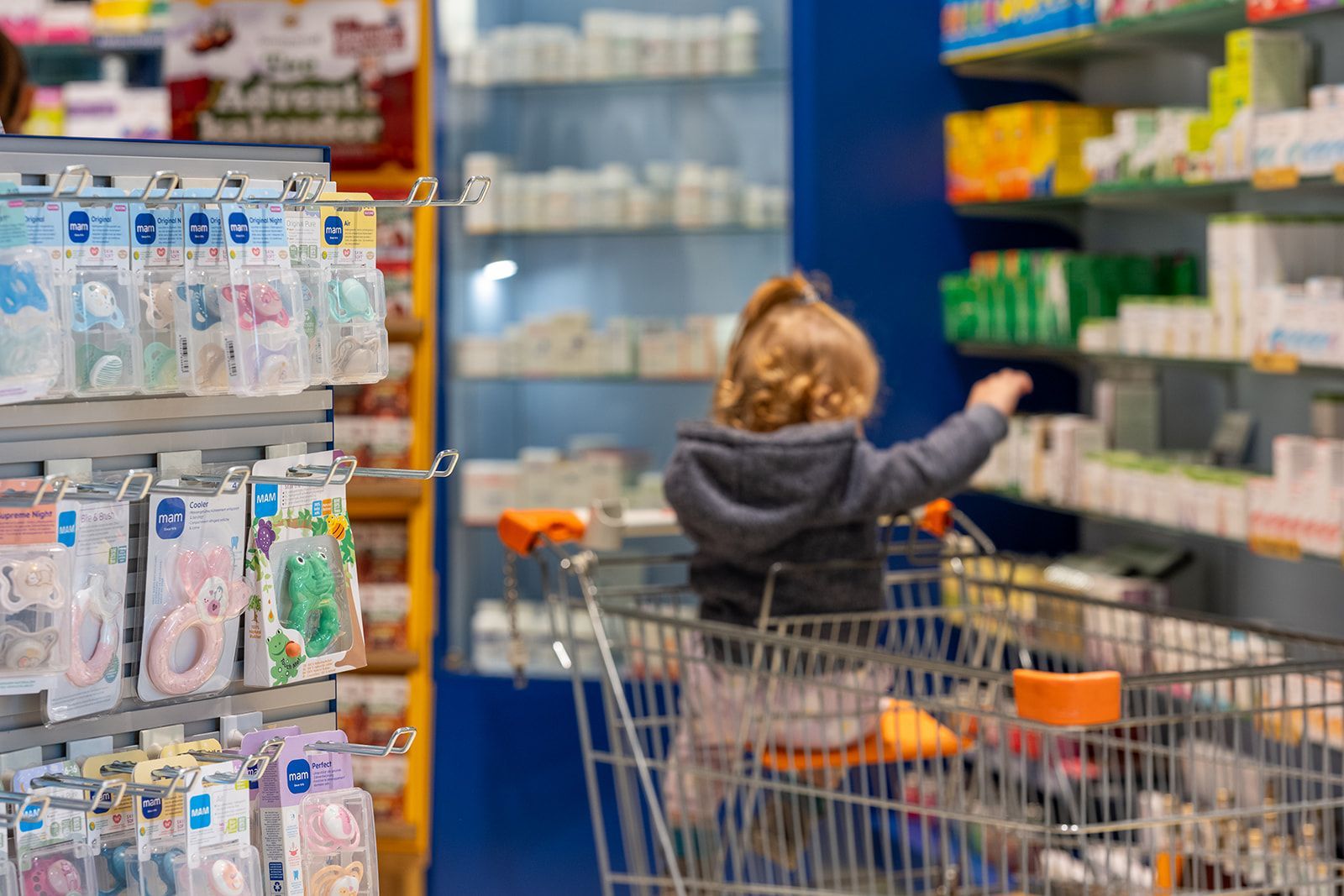 Kind in einem Einkaufswagen greift nach Produkten auf einem Regal in einer Apotheke.