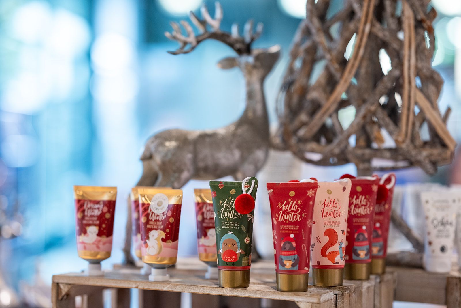 Handcremes mit Weihnachtsmotiven auf einem Holzdisplay, mit einer Rentierstatue und einem dekorativen Baum im Hintergrund.