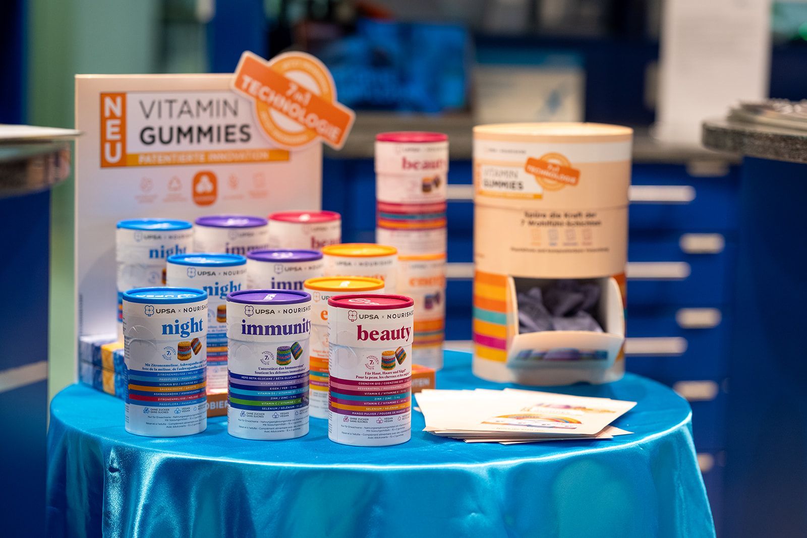 Vitamingummi-Ausstellung auf einem blauen Tisch: mehrere bunte Behälter und ein Werbeschild.