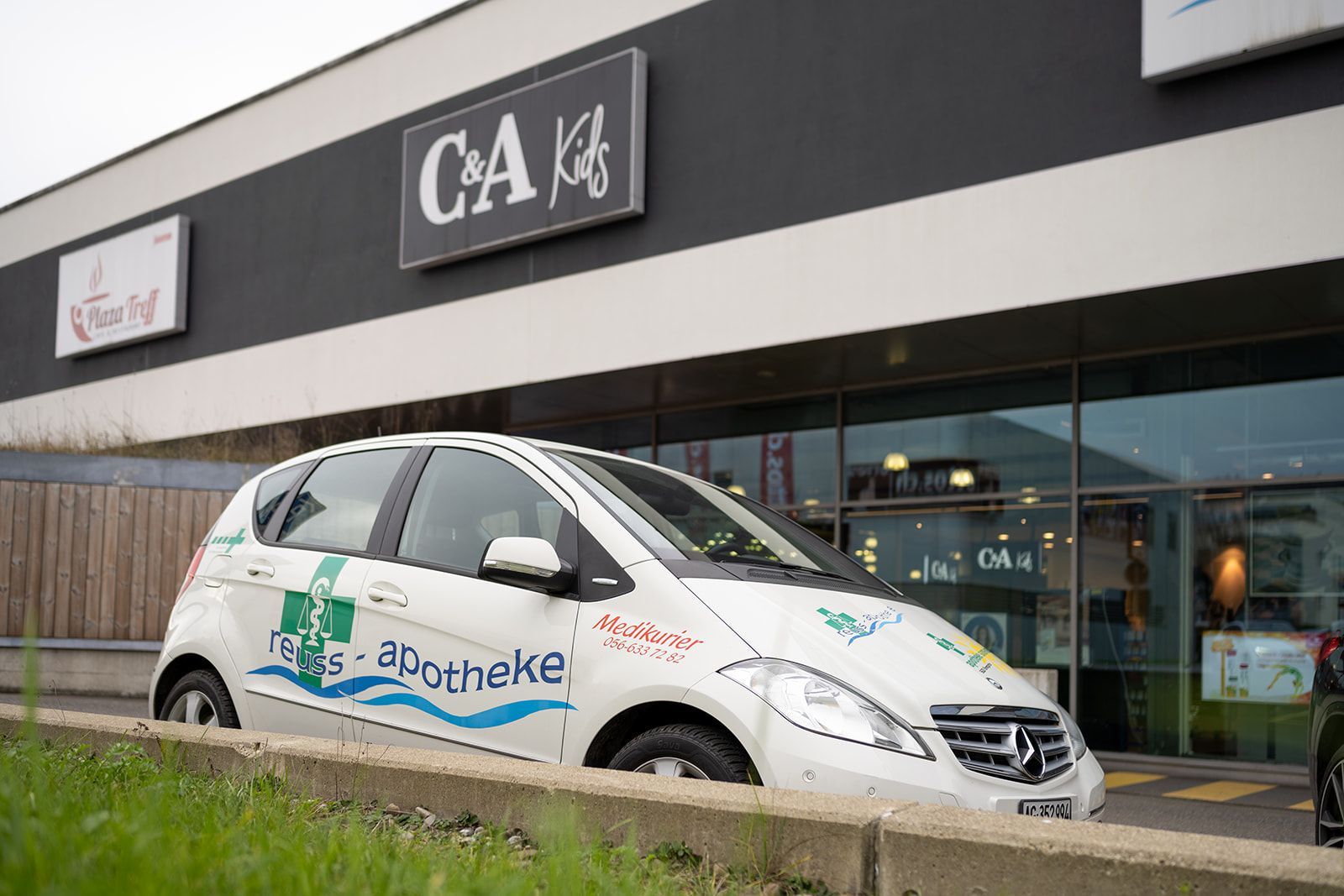 Weißes Auto mit Apotheken-Branding vor einer C&A-Filiale geparkt.
